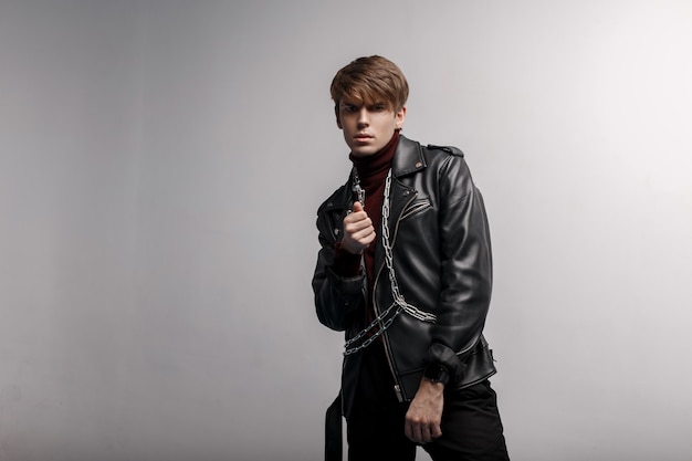 Handsome attractive man in a black trendy jacket in retro style with a hair style in red golf and black trousers with a silver chain, posing in a studio near a white wall. Modern stylish guy