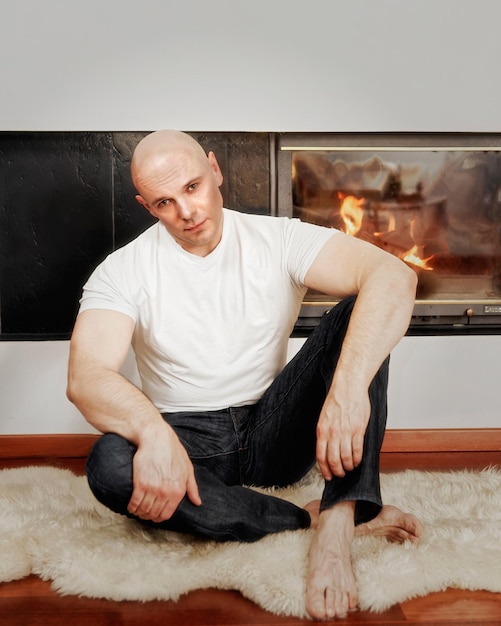 Handsome bald headed man sitting on fur carpet near fireplace. Fire in the fireplace is burning.