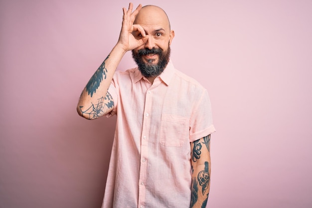 Handsome bald man with beard and tattoo wearing casual shirt over isolated pink background doing ok gesture with hand smiling eye looking through fingers with happy face