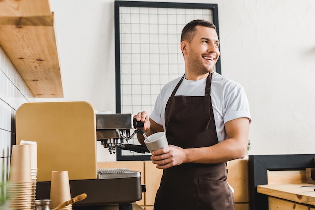 Handsome barista holding cup and making coffee with machine in coffee house