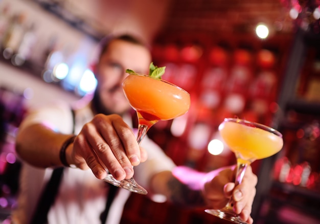 Handsome bartender prepares an orange alcoholic cocktail and smiles on the surface of a bar or a night club