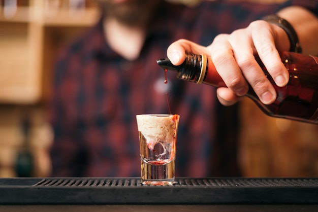 Красивый бородатый бармен готовит коктейль из обезьяньих мозгов в ночном клубе. Селективный фокус на стекле.