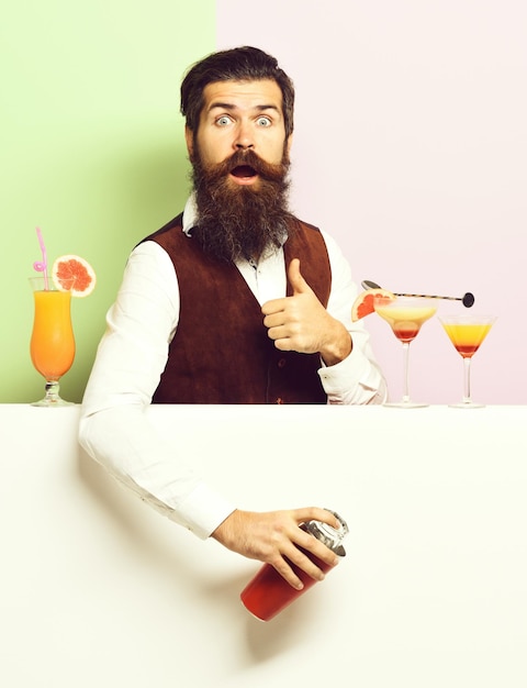 Handsome bearded barman with long beard and mustache has stylish hair on surprised face holding shaker and made alcoholic cocktail in vintage suede leather waistcoat on purple green studio background