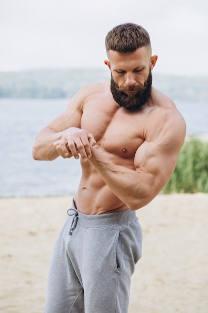 Handsome bearded bodybuilder man with perfect muscular body posing outdoors near lake. Strong man with naked torso standing, looking away.