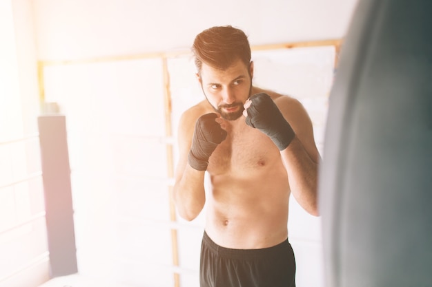 Handsome bearded boxer with bare torso is practicing punches at the fight club