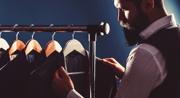 Handsome bearded businessman in classic suit. Businessman in vest, row of suits in shop. It is in the showroom, trying on clothes, posing. Stylish man in a cloth jacket