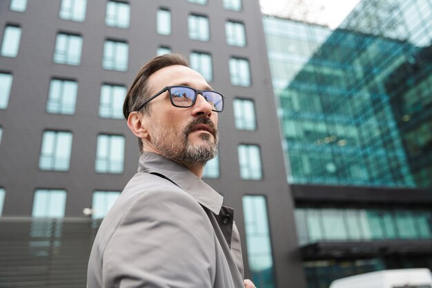 handsome bearded businessman in eyeglasses posing and looking forward near office building in urban area