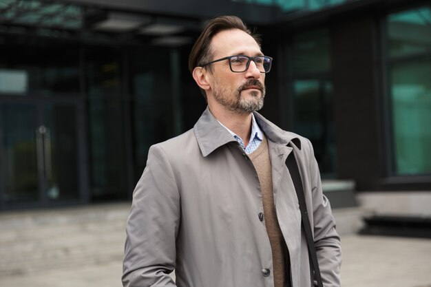 handsome bearded businessman in eyeglasses posing and looking forward near office building in urban area