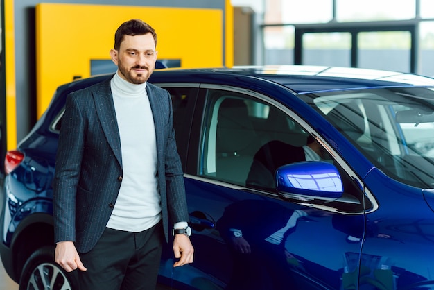 Handsome bearded buyer in casual wear in dealership, guy looks on camera while standing near car with crosed arms