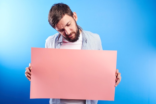 Handsome bearded caucasian man holding blank pink poster board in hands with copy space for text or advertising inscription isolated in blue studio