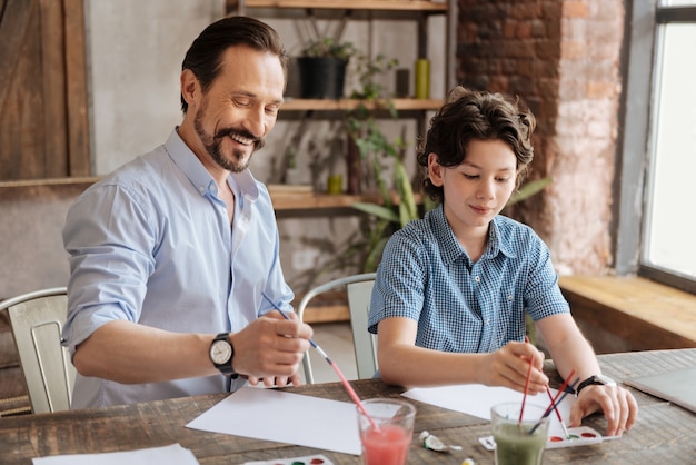 Handsome bearded father and his charming son sitting at the table in the living room and enjoying themselves while starting to paint new pictures