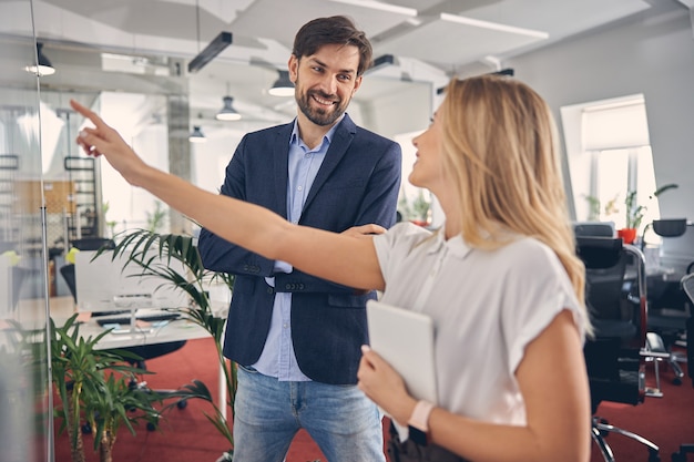 Handsome bearded gentleman looking at female colleague and smiling while lady pointing at whiteboard