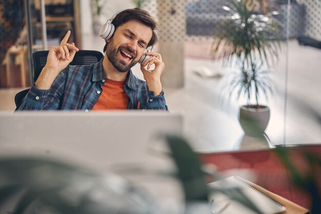 Handsome bearded gentleman in wireless headphones enjoying favorite songs while sitting at the table in office