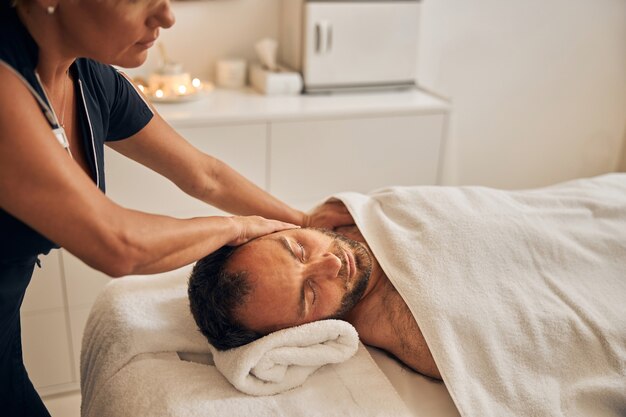 Photo handsome bearded gentleman with closed eyes receiving therapeutic massage while lying on massage table under white blanket