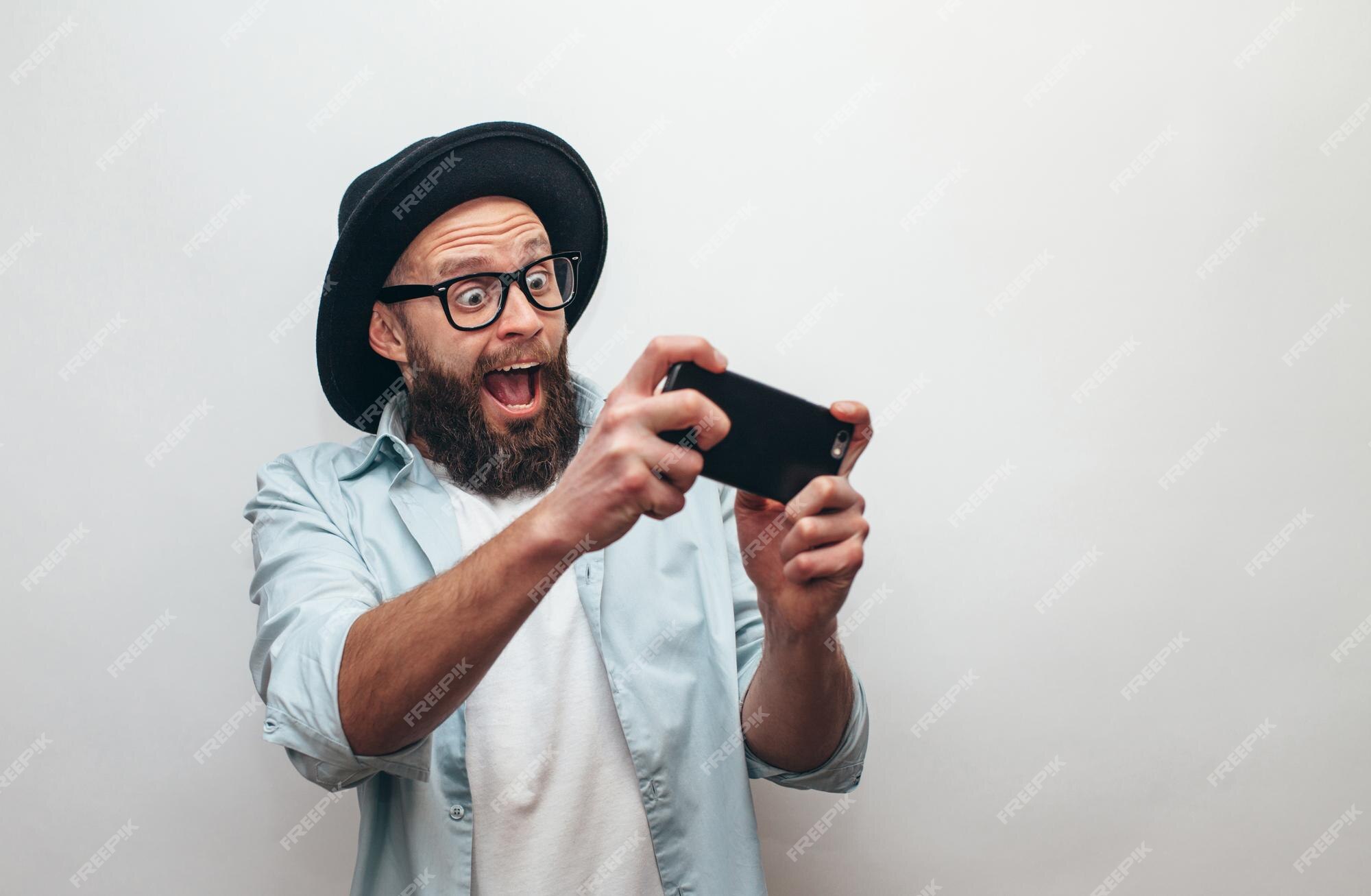 Premium Photo  Handsome bearded guy holds smartphone and plays online games  joyful man wearing a black stylish hat and a blue shirt crazy emotions