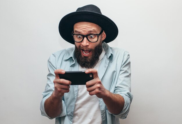 Premium Photo  Handsome bearded guy holds smartphone and plays online games  joyful man wearing a black stylish hat and a blue shirt crazy emotions