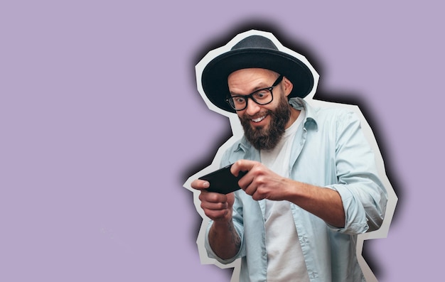 Handsome bearded guy holds smartphone and plays online games Joyful man wearing a black stylish hat and a blue shirt Crazy emotions