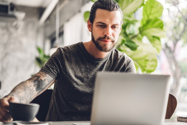 Foto uso barbuto bello dell'uomo dei pantaloni a vita bassa ed esaminare computer portatile con caffè alla tavola in caffè concetto di tecnologia e di comunicazione