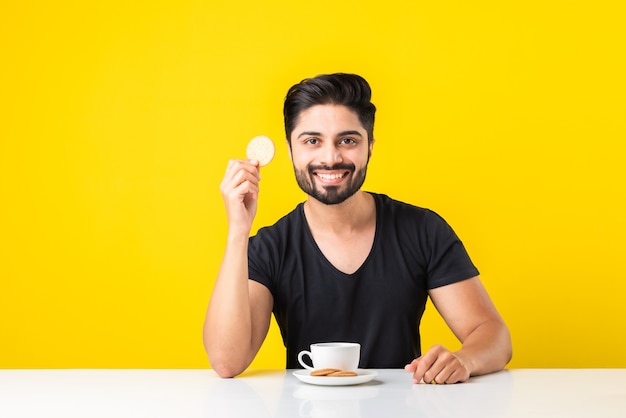 Handsome bearded Indian man eating healthy whole grain biscuits dipped in coffee of Tea or chai in a cup, sitting at table against yellow background