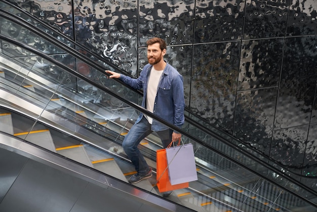 Красивый бородатый мужчина с сумками для покупок поднимается на эскалаторе торгового центра