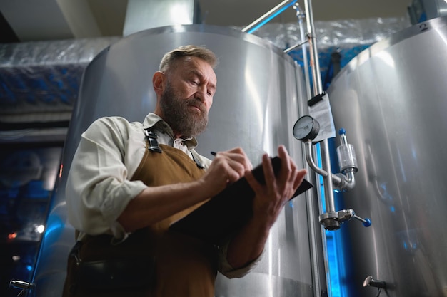 Handsome bearded man brewer inside modern beer factory around steel tanks. Mature male worker making notes about technological process