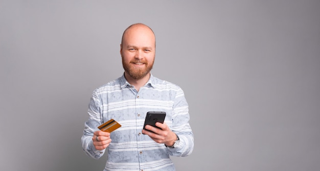 Handsome bearded man in casual using debit card and smartphone fro online payments over grey background