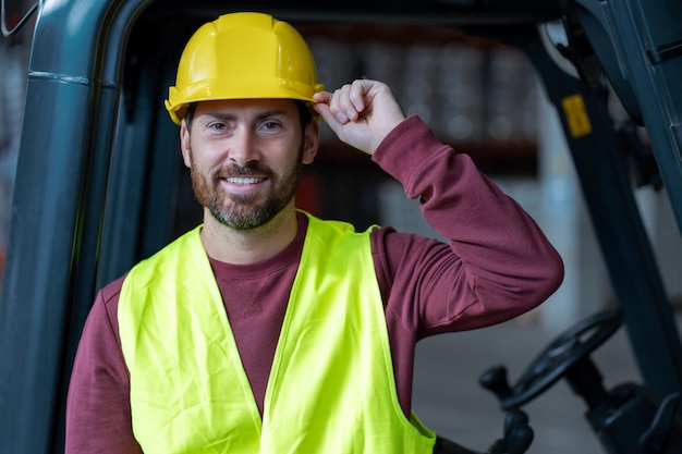 Handsome bearded man driver wearing helmet and workwear standing near forklift looking at camera