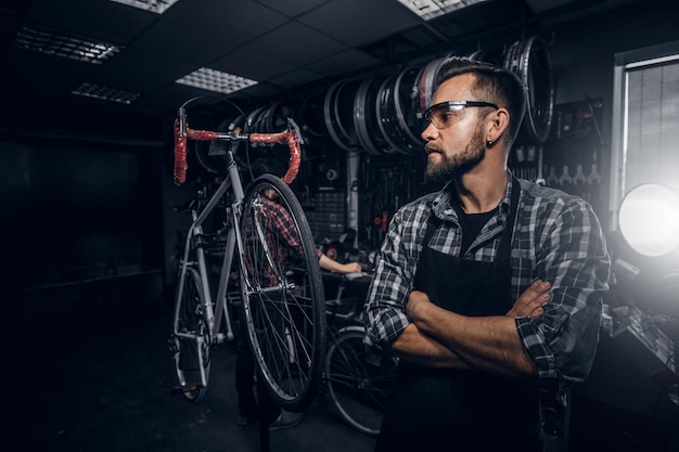 Photo handsome bearded man in glasses is standing near fixed bicycle at his own workshop.