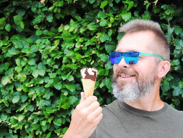 A handsome bearded man in green sunglasses holds an ice cream cone with chocolate in his hands and smiles