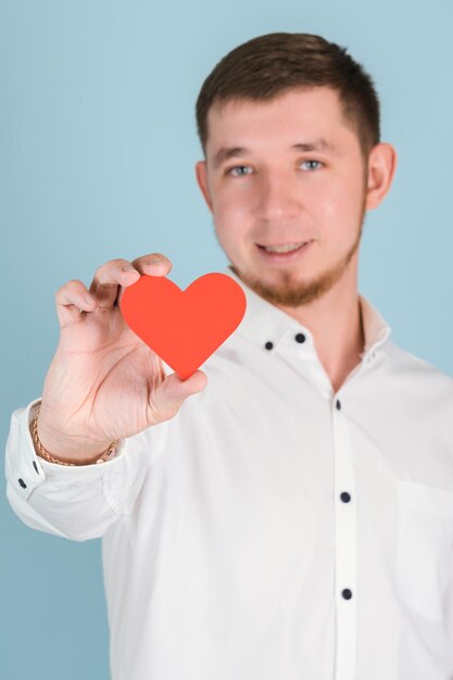 A handsome bearded man holds a red heart symbol at arm's length standing on a blue background, selective focus