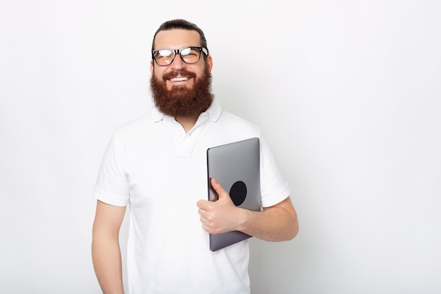 L'uomo barbuto bello tiene in mano un computer portatile chiuso su sfondo bianco.