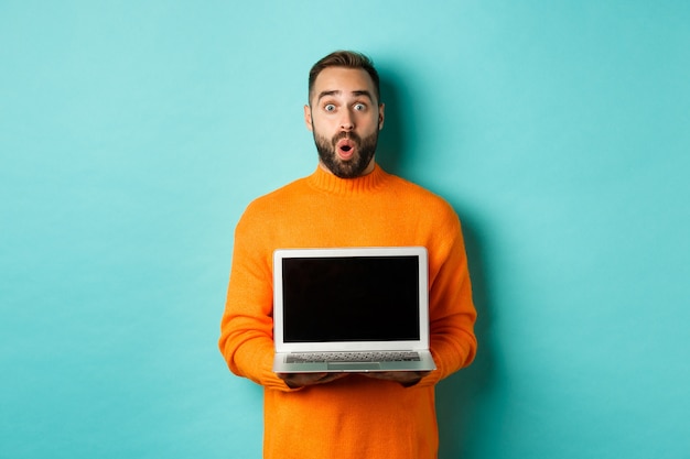 Handsome bearded man in orange sweater showing laptop screen, demonstrating promo, standing over light blue background.