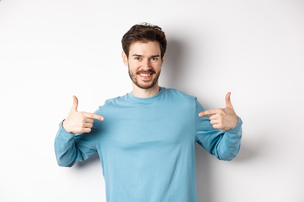 Photo handsome bearded man pointing fingers at center, self-promoting and looking confident, standing over white background, smiling at camera