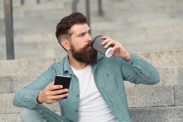 携帯電話とコーヒーカップをリラックスしてハンサムなひげを生やした男。オンライン生活を送る。ソーシャルネットワーク。現代の生活オンラインコミュニケーション。スマートフォンの都市空間の背景を持つ男。現代のテクノロジー。