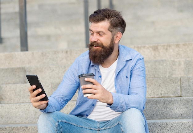 Handsome bearded man relaxing mobile phone and coffee cup Modern technology Living online life Social networks Modern life online communication Guy with smartphone urban space background