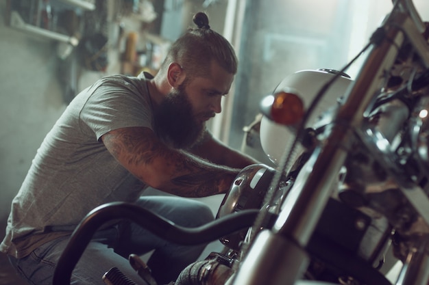 ハンサムなひげを生やした男がガレージで彼のバイクを修理します。ジーンズとTシャツを着た男性