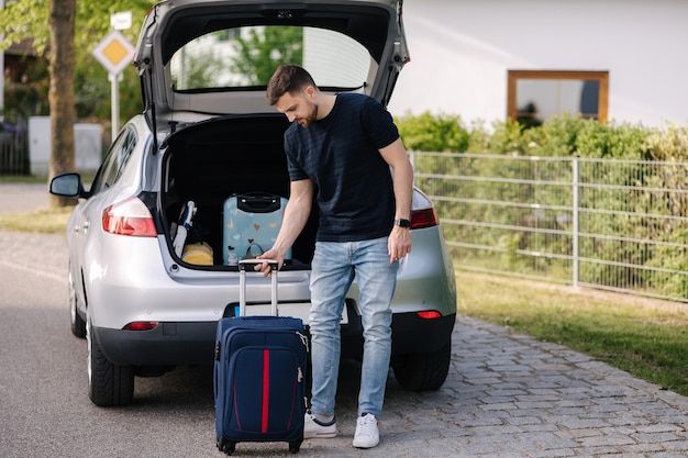 Handsome bearded man stand with suitcase in front of open car trunk Travel road trip Holiday summer vacation concept