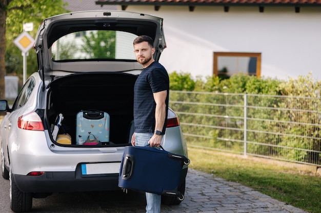 Handsome bearded man stand with suitcase in front of open car trunk travel road trip holiday summer