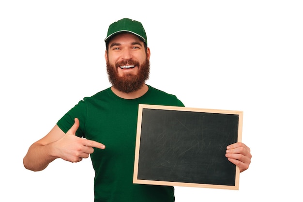 Handsome bearded man wearing green t shirt and cap pointing at black board