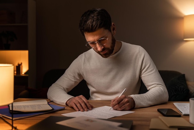 Handsome bearded man wearing white sweater writing letter at home during cozy evening. People concept. Stock photo