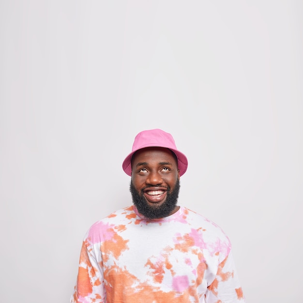 Handsome bearded man with happy smile looks above being in good mood wears panama and casual t shirt poses against white wall