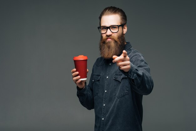 スタイリッシュな髪のひげと深刻な顔に口ひげを生やしたハンサムなひげを生やした男がカメラを保持しているカップまたは灰色の空間でお茶やコーヒーを飲むマグカップを指して