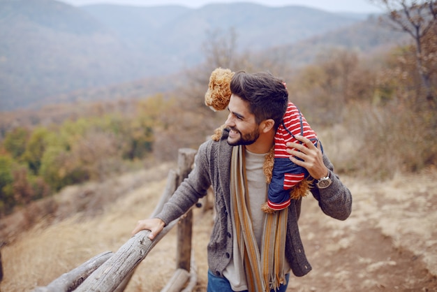 Photo handsome bearded mixed race man dressed casual walking in nature at fall and carrying his apricot poodle on shoulders.