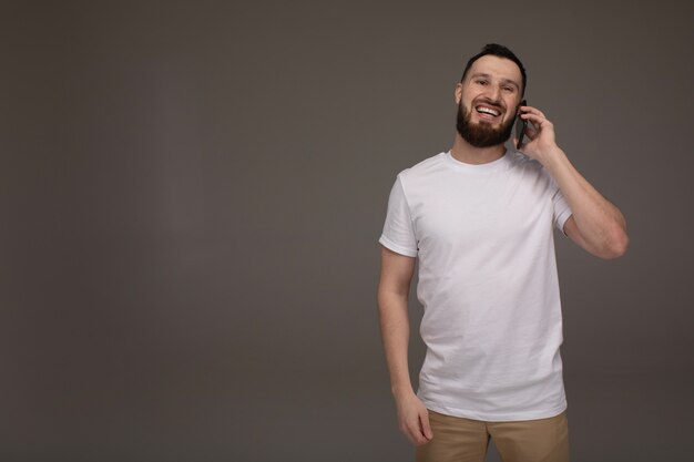 Handsome bearded model is posing with phone in hands over grey background.