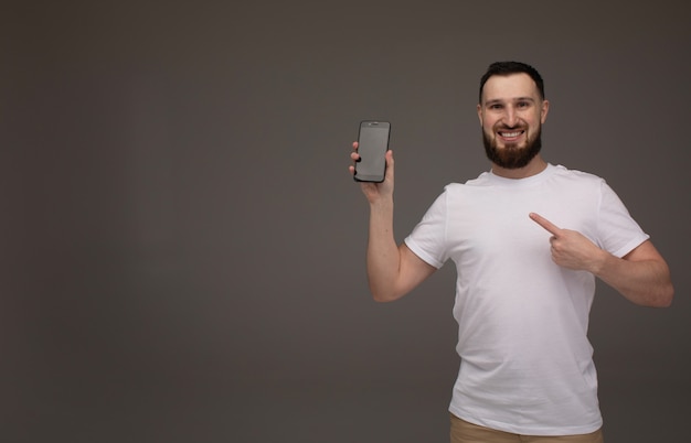 Handsome bearded model is posing with phone in hands over grey background.