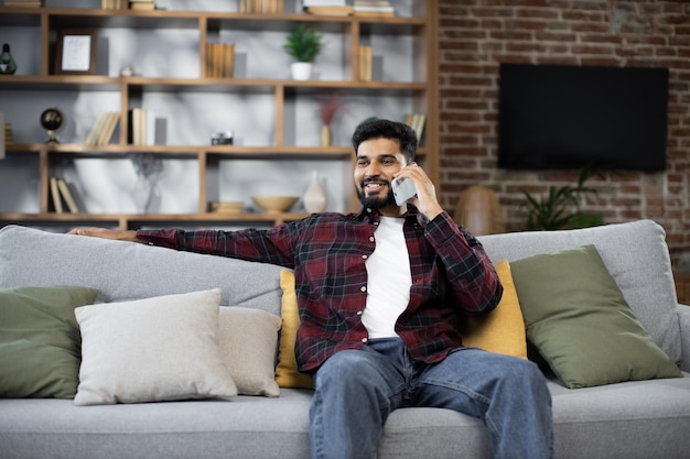 Handsome bearded positive man sitting on couch and calling to\
his friend using smartphone indoors