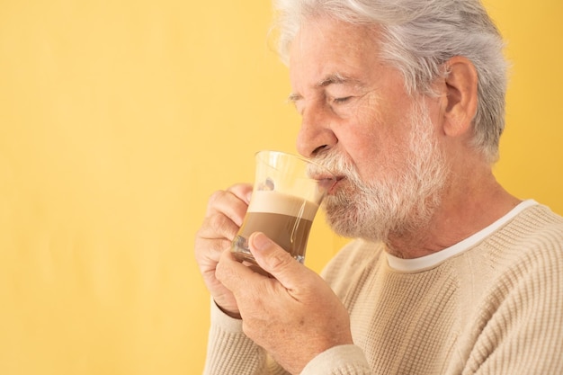 Un bell'uomo anziano barbuto che beve caffè e cappuccino da un bicchiere di vetro, in piedi su sfondo giallo