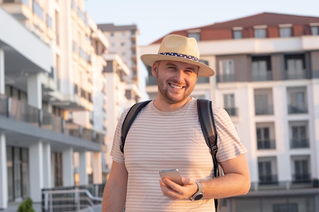 Foto un bel mantourista sorridente con la barba che guarda la macchina fotografica, che indossa un cappello solare, uno zaino che tiene in mano uno smartphone.