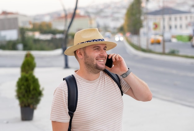 Foto bel manturista sorridente con la barba che indossa uno zaino con cappello da sole che chiama utilizzando lo smartphone in città all'aperto felice ragazzo caucasico che parla al telefono in viaggio