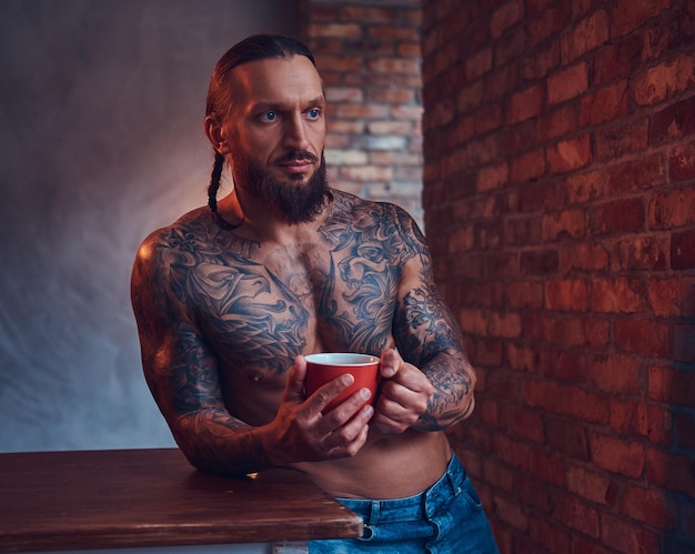 Handsome bearded tattoed male with a stylish haircut and muscular body, drinks coffee, leaning on a table.